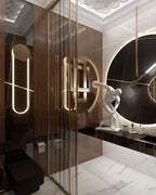 Inside a marble and glass washroom, a Clérachic wall light with a long LED loop glows warm beside mirrored panels and a round black mirror.