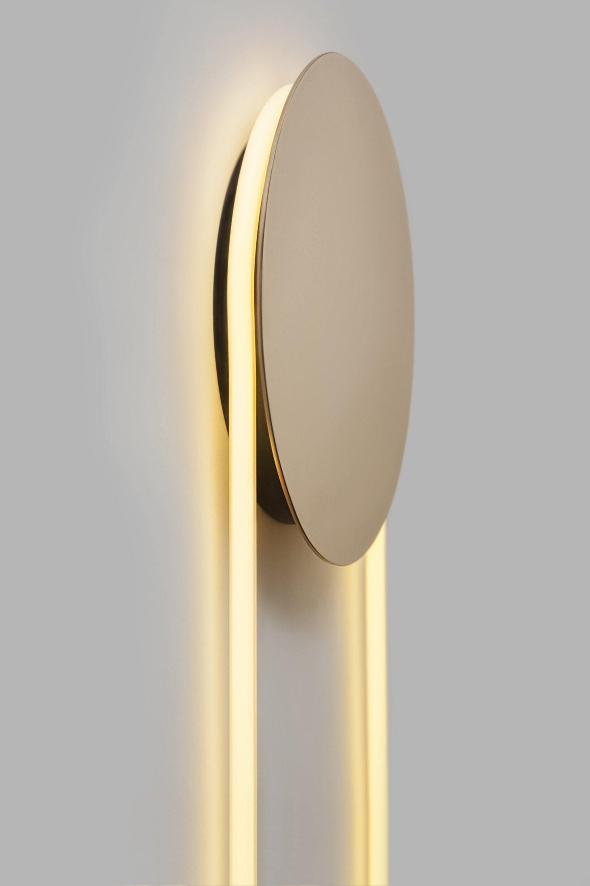 Angled close view of the polished gold disc and glowing LED loop on this Clérachic wall light, lit warm on a grey wall.
