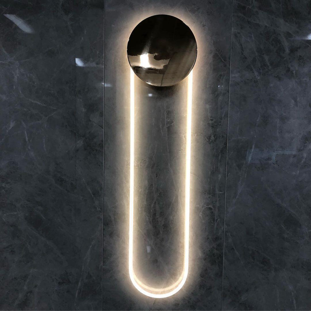 This Clérachic wall light on a dark marble-effect wall, its polished gold disc reflecting the room and the LED loop glowing warm white.