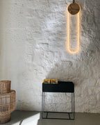 Against a rough white plaster wall, a Clérachic wall light lights up a warm loop above a black plant stand and woven baskets.