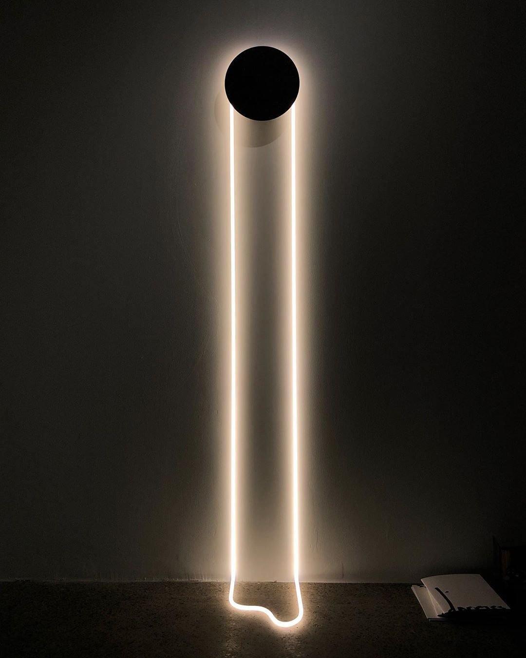 On a dark wall, this Clérachic wall light shows a black disc and a bright white LED loop whose tail curls on the floor below.