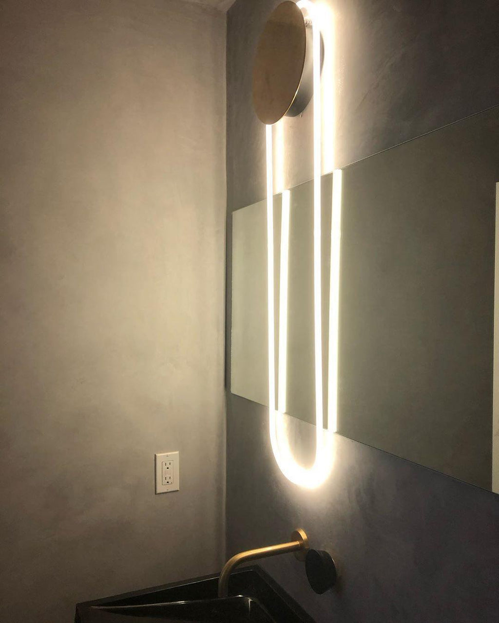 Above a washbasin with a brass tap, a Clérachic wall light lights a long LED loop beside a wide mirror on a grey plaster wall.