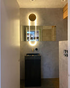 In a small washroom, a Clérachic wall light with a gold disc and a lit LED loop sits above a black basin and a brass wall tap.