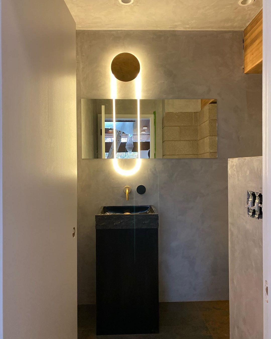 In a small washroom, a Clérachic wall light with a gold disc and a lit LED loop sits above a black basin and a brass wall tap.