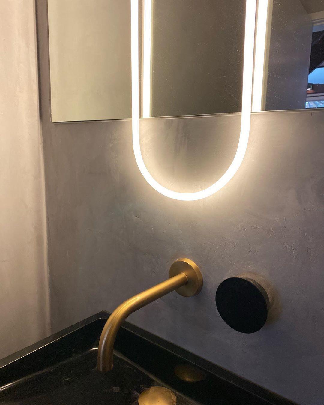 Close view of the lower curve of the lit LED loop on this Clérachic wall light, above a brass tap and a black basin edge.