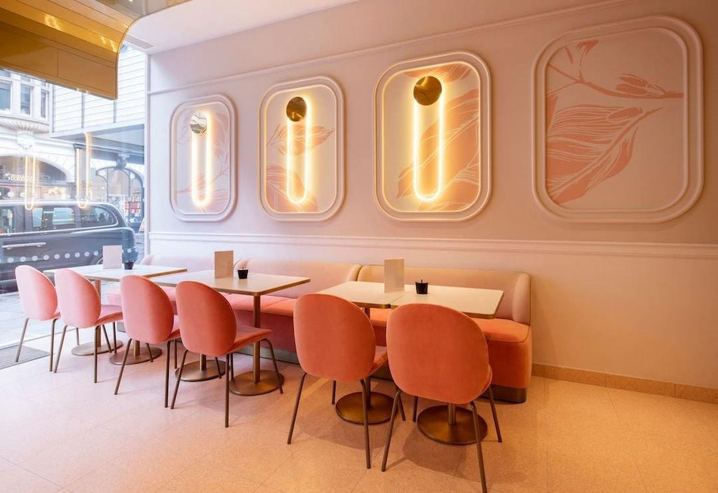 Wide view of a pale pink café, with Clérachic wall lights glowing in recessed panels above pink chairs and a long banquette.