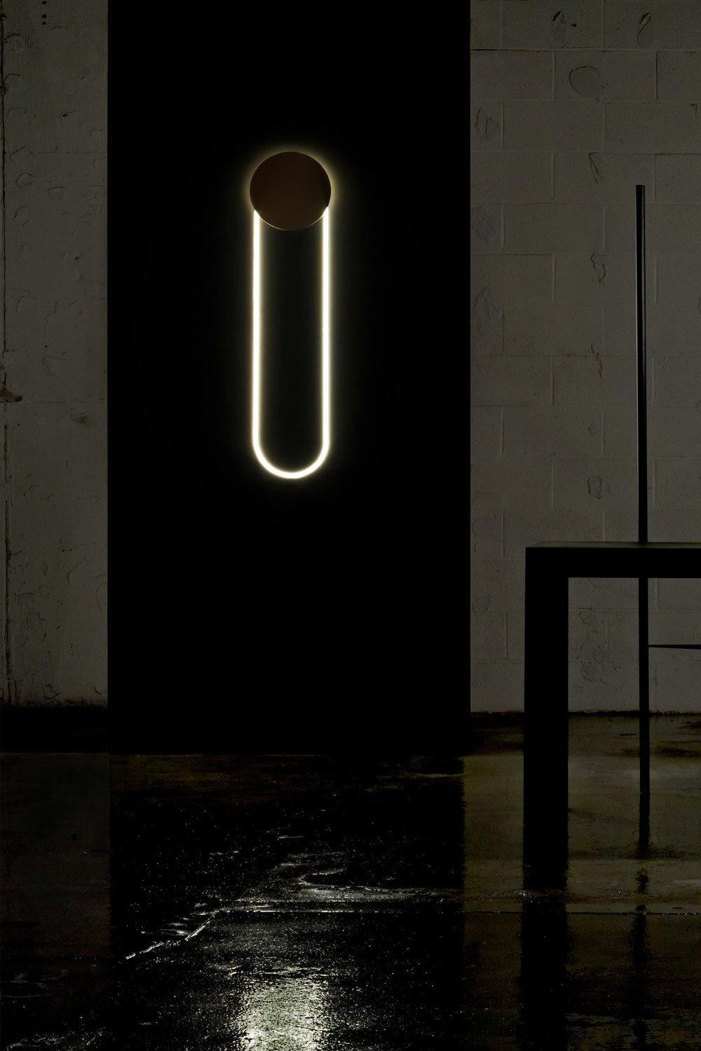 In a dark, wet-floored space, a Clérachic wall light shows a gold disc and a bright white LED loop against a black wall.