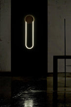 In a dark, wet-floored space, a Clérachic wall light shows a gold disc and a bright white LED loop against a black wall.
