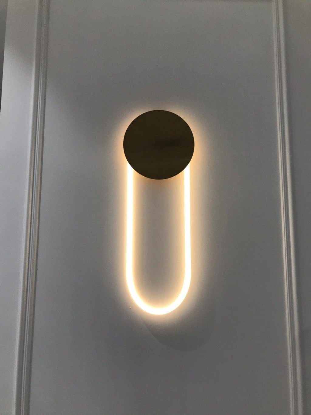 On a pale grey wall with moulded panels, a Clérachic wall light shows a bronze disc and a warm LED loop in an even glow.