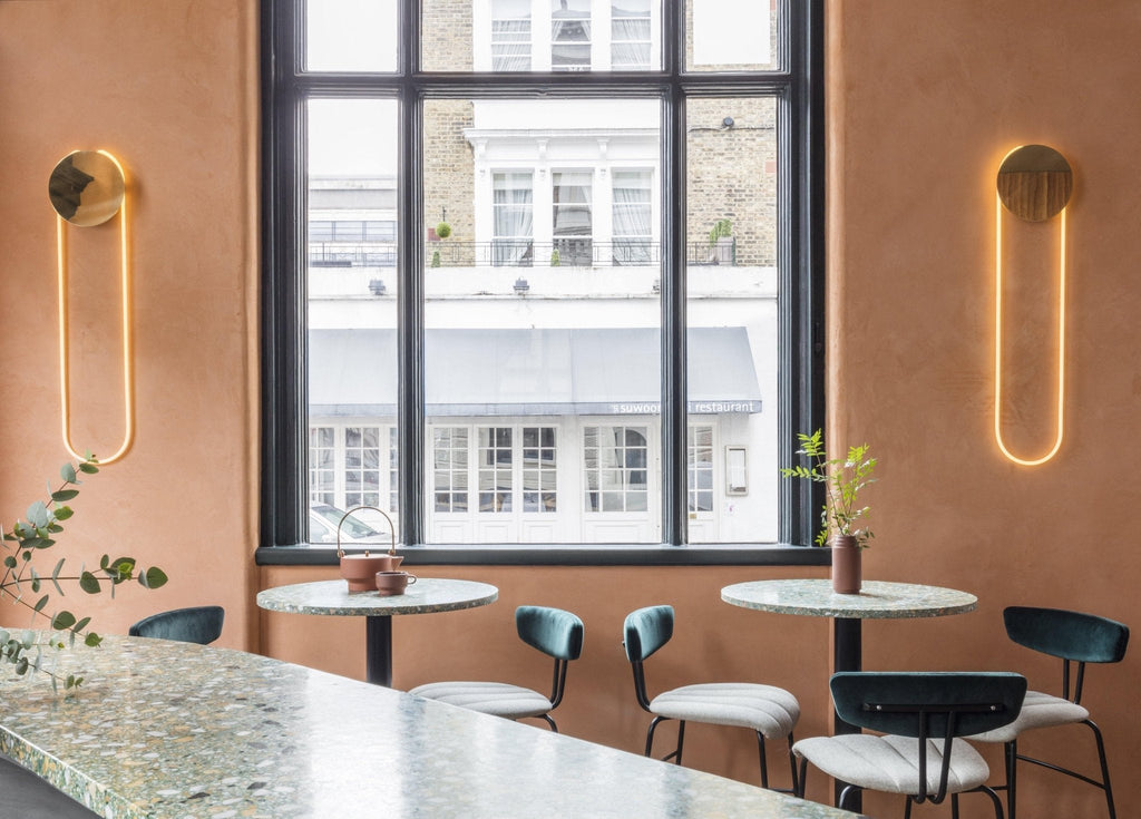 Two Clérachic wall lights with gold discs and warm LED loops flank a tall black-framed window above round tables and dark chairs.