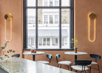 Two Clérachic wall lights with gold discs and warm LED loops flank a tall black-framed window above round tables and dark chairs.
