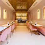 Looking down a pale pink café, Clérachic wall lights glow in the arched panels along both walls above pink chairs and a counter.
