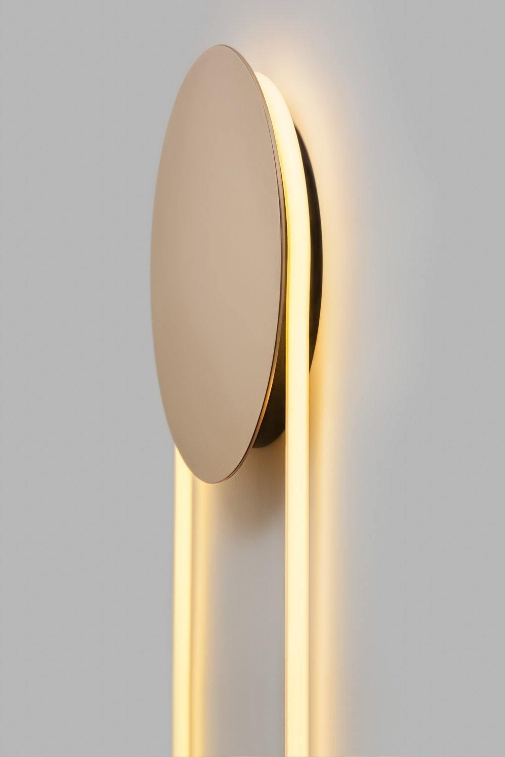 Angled view of the polished gold disc of this Clérachic wall light, standing off a grey wall with a warm halo around it.