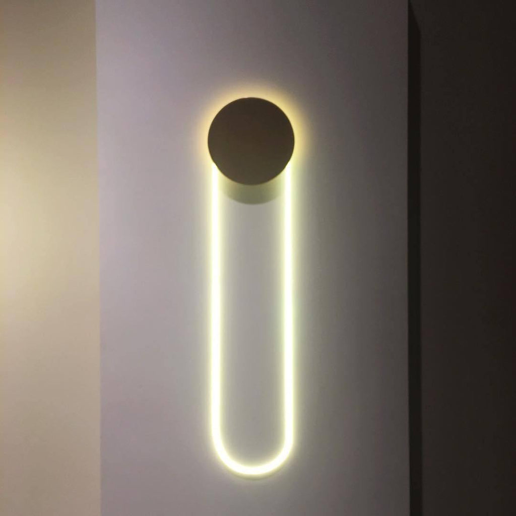 A Clérachic wall light on a grey wall between dark panels, its dark disc above a long LED loop glowing a cool, clean white.