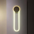 A Clérachic wall light on a grey wall between dark panels, its dark disc above a long LED loop glowing a cool, clean white.