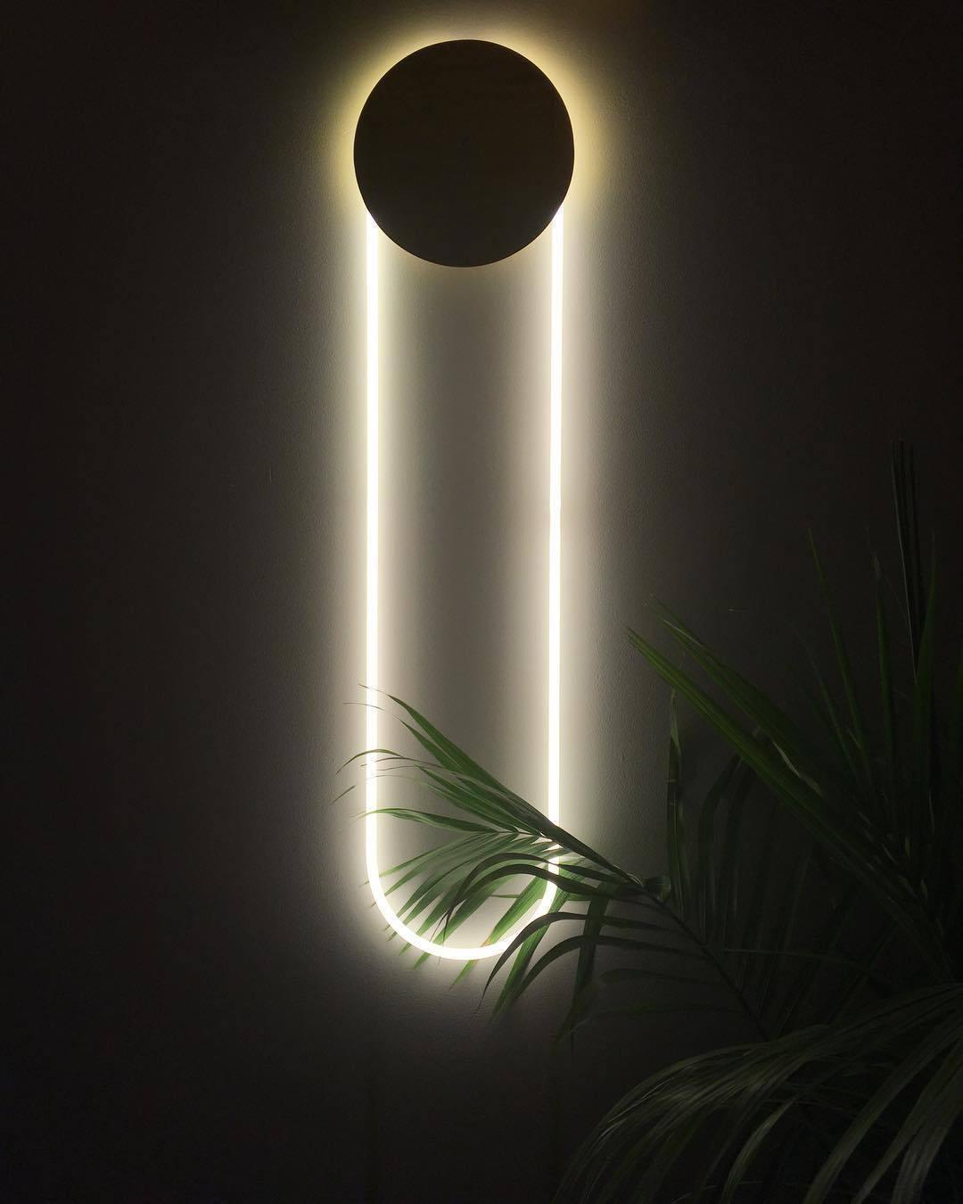 This Clérachic wall light on a black wall, with a dark disc and a white LED loop lit bright, and the tip of a palm in front.