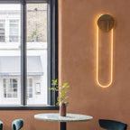A Clérachic wall light with a wooden-look disc and a warm LED loop on a terracotta wall, beside a window and a small plant on a round table.