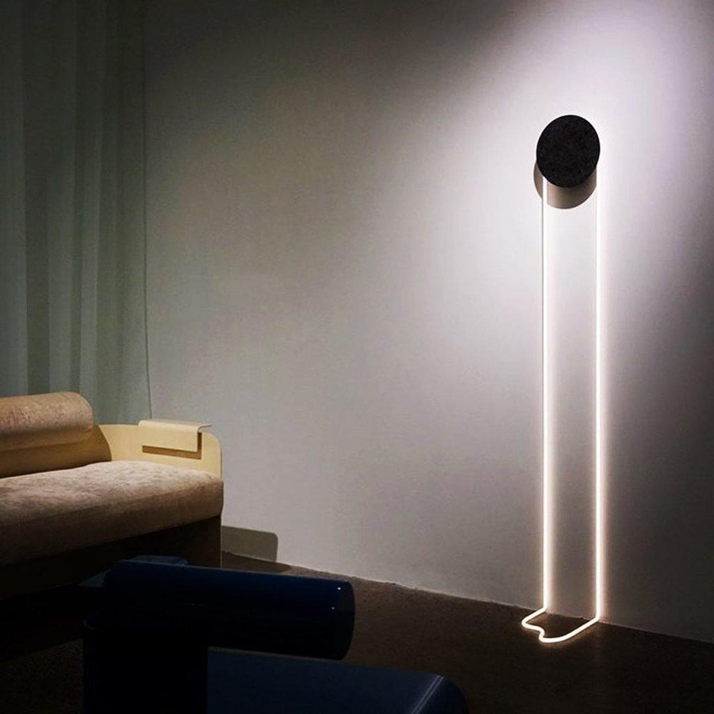This Clérachic wall light with a long white LED loop on a dark wall, so tall that its tail reaches the floor beside a sofa.