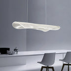 Angled view of a Clérachic pendant light with a rippled clear shade, above a long white table with grey chairs on a blue-grey wall.