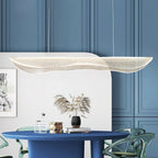 A Clérachic pendant light with a rippled shade above a round blue dining table with green glass chairs, against a blue panelled wall.