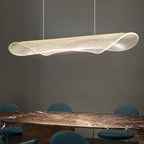 Angled view of a Clérachic pendant light with a curled, clear acrylic shade above a dark marble table and teal chairs.