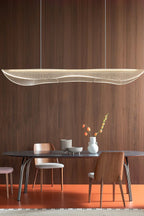 Clérachic pendant with a lit, wavy acrylic shade hung in front of a walnut wall, above a dark oval table on an orange floor.
