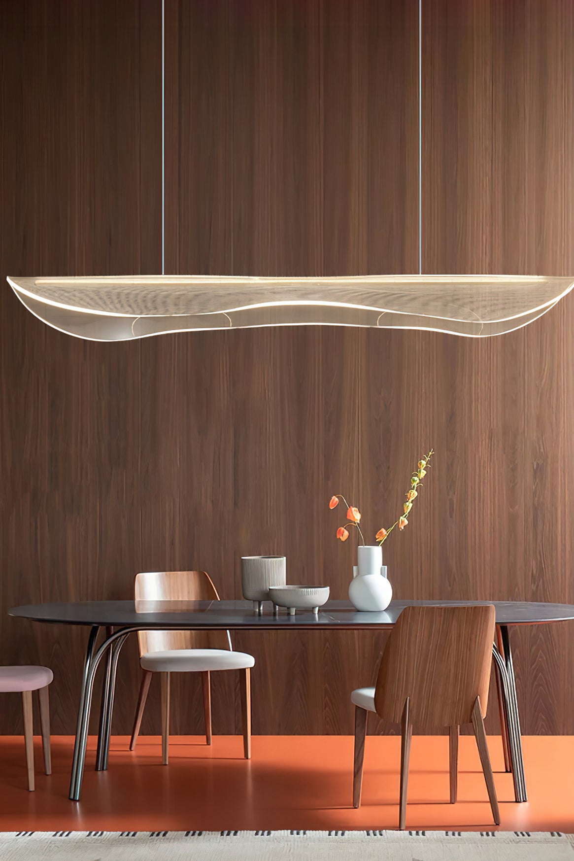 Clérachic pendant with a lit, wavy acrylic shade hung in front of a walnut wall, above a dark oval table on an orange floor.