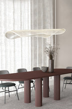 Clérachic pendant light with a curled clear shade above a long burgundy dining table with fluted legs and grey chairs, by sheer curtains.
