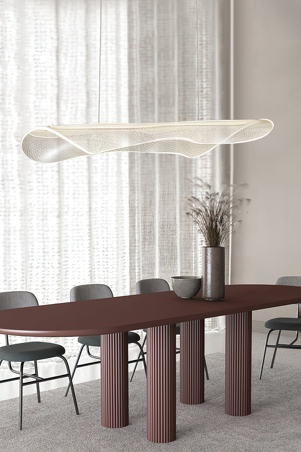 Clérachic pendant light with a curled clear shade above a long burgundy dining table with fluted legs and grey chairs, by sheer curtains.