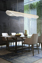 A Clérachic pendant light with a curled clear shade hung above a wooden dining table with a fruit bowl, beside a window with a green wall.