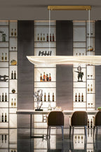 Clérachic linear pendant light with a gold canopy above a marble table, in front of shelves of bottles and glasses with brown dining chairs.