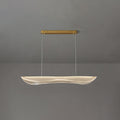 A Clérachic pendant light lit at a shorter drop, the clear wavy acrylic shade glowing under a slim gold canopy on a grey wall.
