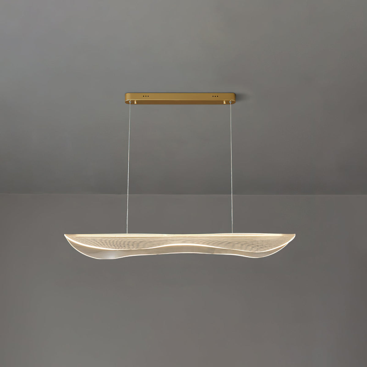 A Clérachic pendant light lit at a shorter drop, the clear wavy acrylic shade glowing under a slim gold canopy on a grey wall.