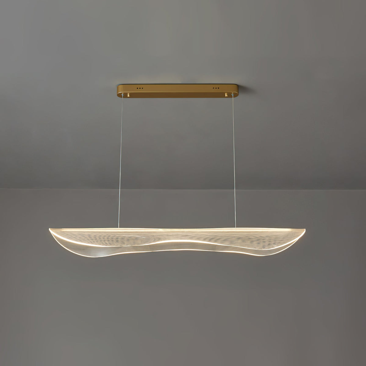 Clérachic linear pendant with a warm light along the rim of its wavy acrylic shade, hung under a gold canopy against grey.