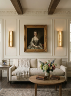 Pair of slim brass Clérachic alabaster sconces flanking a gilt-framed portrait, above a cream sofa in a panelled room.