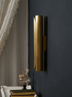 Tall curved brass Clérachic wall sconce on a deep navy wall, seen from the side beside a curtain and a small table of books.