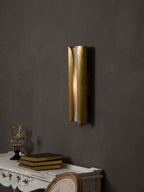 Clérachic brass sconce with a lit alabaster centre on a dark wall, above a carved white console with books and a glass vase.