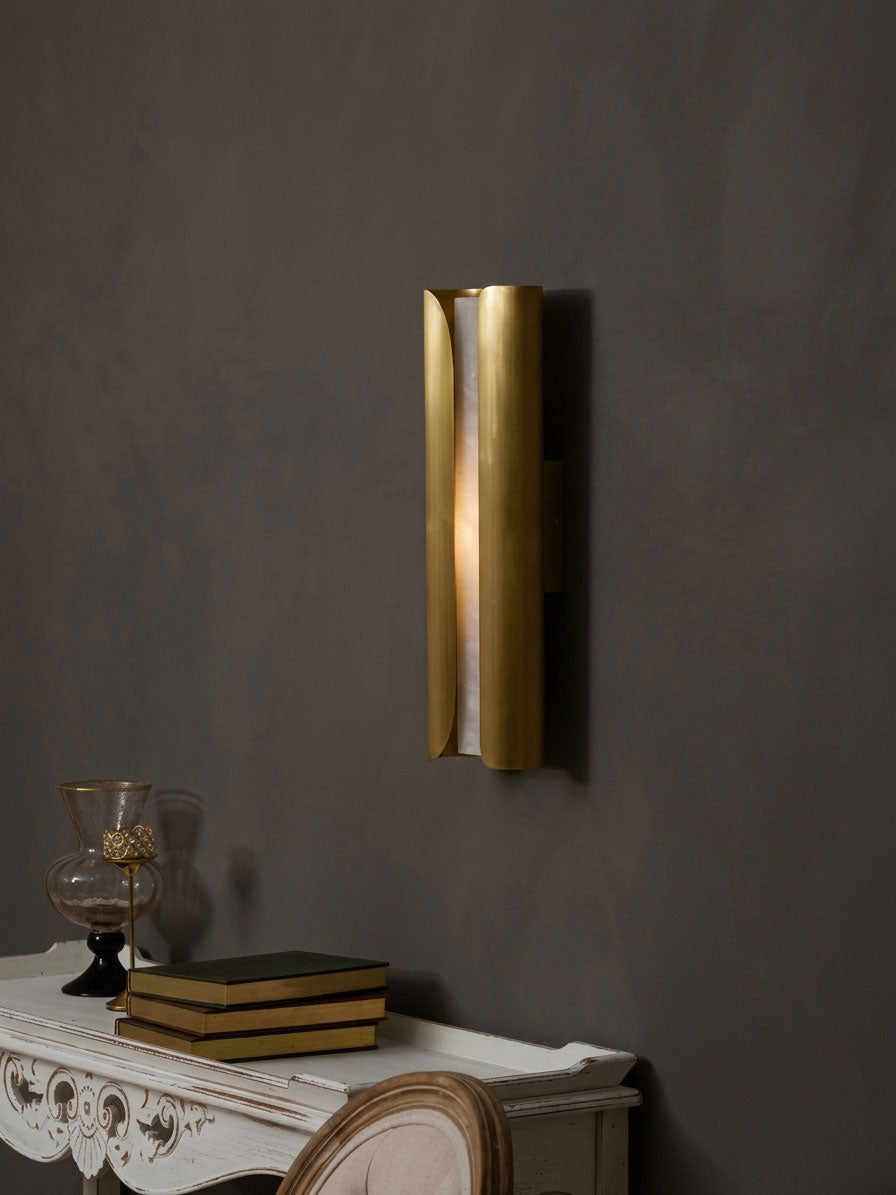 Clérachic brass sconce with a lit alabaster centre on a dark wall, above a carved white console with books and a glass vase.