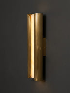 Side view of a curved brass Clérachic wall sconce, its folded metal panel guiding warm light along a dark wall.