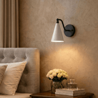 Clérachic matte black cone-shade wall sconce lit above a bedside table with white roses and a tufted headboard.