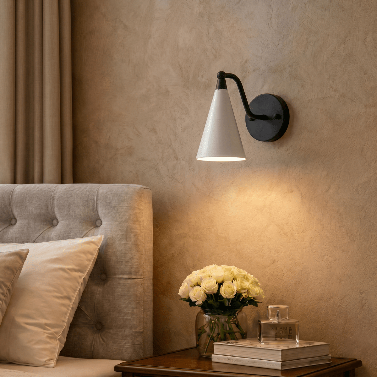 Clérachic matte black cone-shade wall sconce lit above a bedside table with white roses and a tufted headboard.