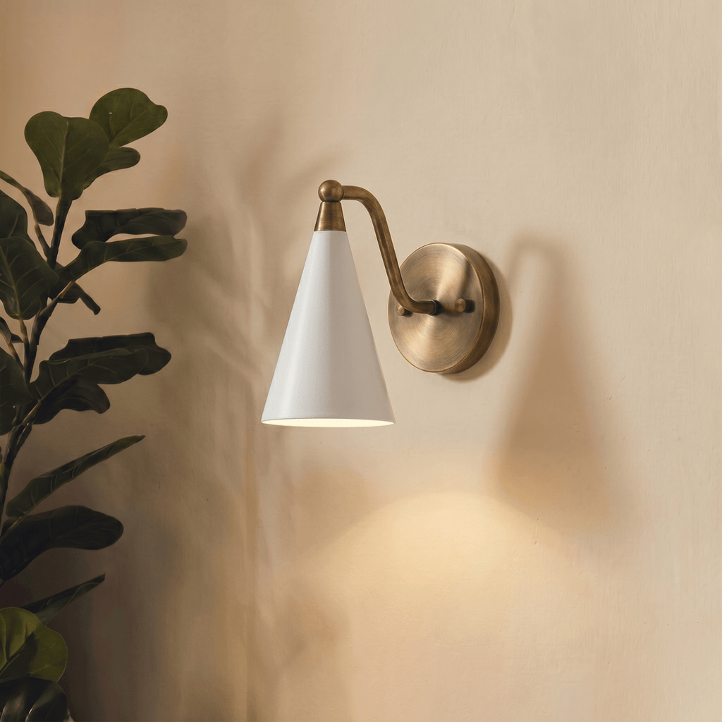 Clérachic vintage brass cone-shade wall sconce lit on a plain wall beside a leafy fiddle-leaf fig plant.