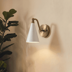 Clérachic vintage brass cone-shade wall sconce lit on a plain wall beside a leafy fiddle-leaf fig plant.