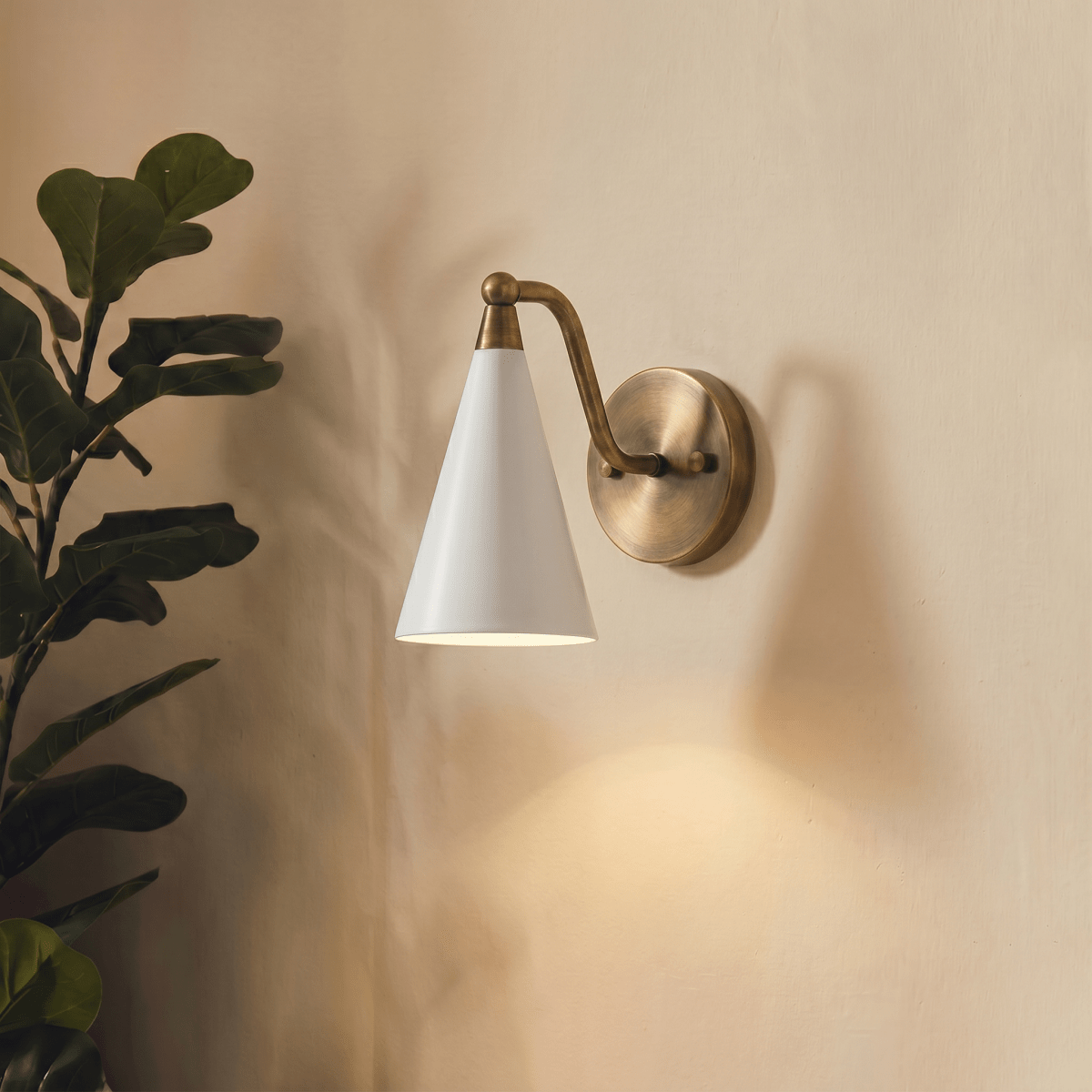 Clérachic vintage brass cone-shade wall sconce lit on a plain wall beside a leafy fiddle-leaf fig plant.