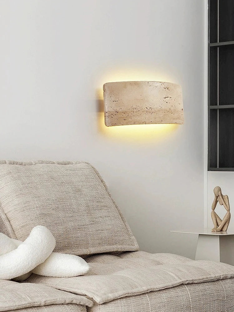 Clérachic travertine wall sconce mounted above a linen sofa in a living room, casting a soft halo of light across the wall behind it.