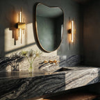 A pair of gold Clérachic wall sconces flanks a curved mirror above a veined marble vanity, lighting the counter below.