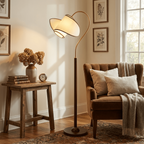 A Clérachic arched floor lamp beside a brown armchair and a rustic stool, with dried flowers in the corner of a warm room.