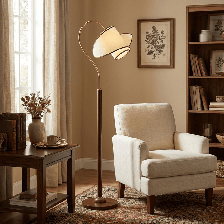 A Clérachic arched floor lamp stands beside a cream armchair, with a bookcase, a botanical print and a dark side table.