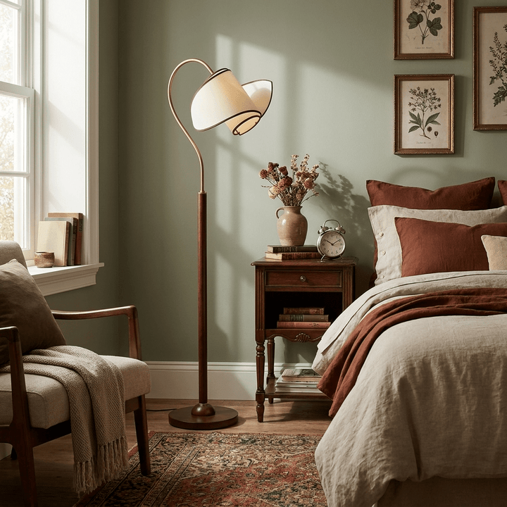 A Clérachic floor lamp lights a sage-green bedroom, beside a bed with rust-coloured bedding and a small wood nightstand.