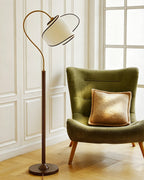 Beside a green velvet chair in front of white panelled walls stands a Clérachic floor lamp with a curved brass arm.